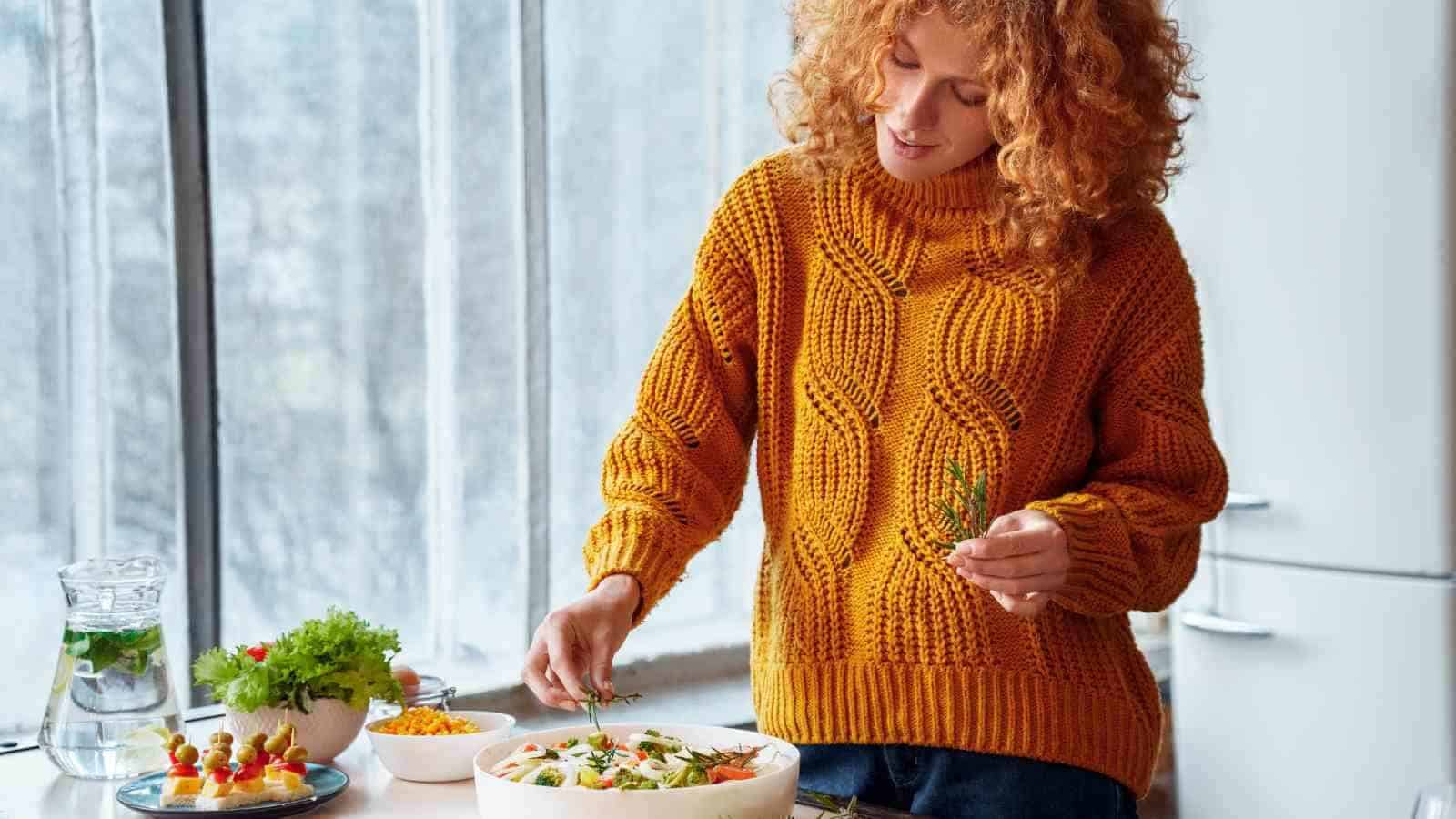 A person in a mustard yellow sweater prepares a salad in a bright kitchen, holding fresh herbs with various ingredients on the counter.