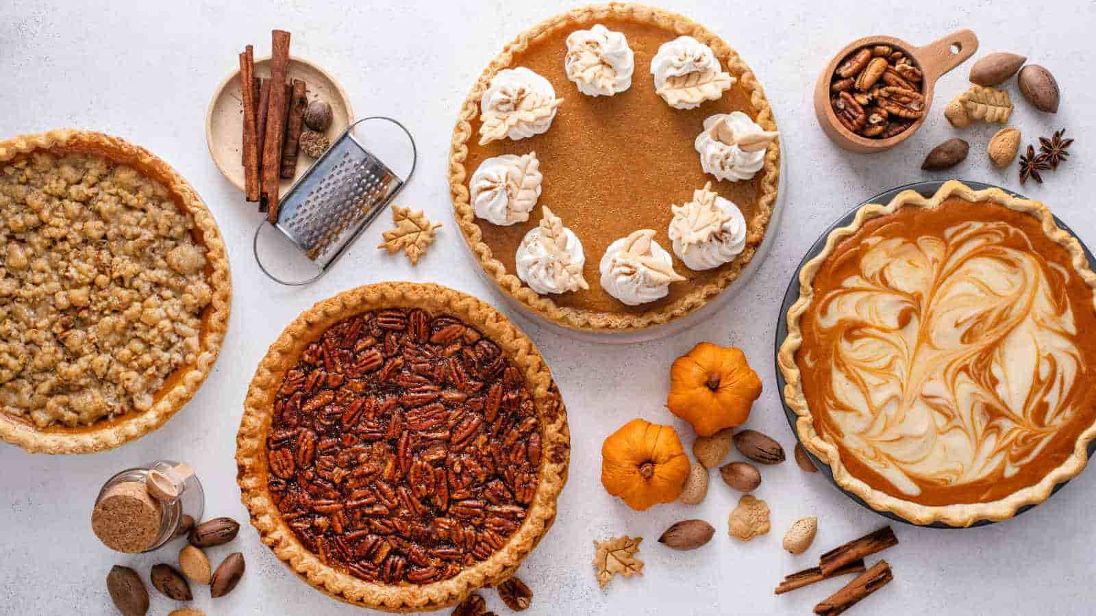 Four pies, including pecan, pumpkin with whipped cream, crumble-topped, and swirled filling, displayed on a white surface with assorted nuts, spices, and small pumpkins.