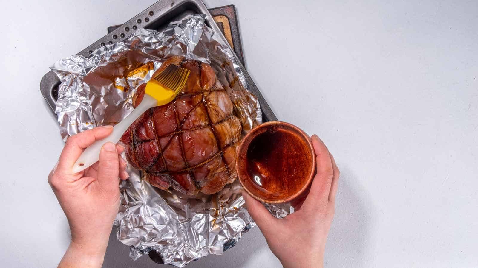 A person brushes glaze onto a cooked ham wrapped in foil on a baking tray, holding a small bowl of glaze in the other hand.