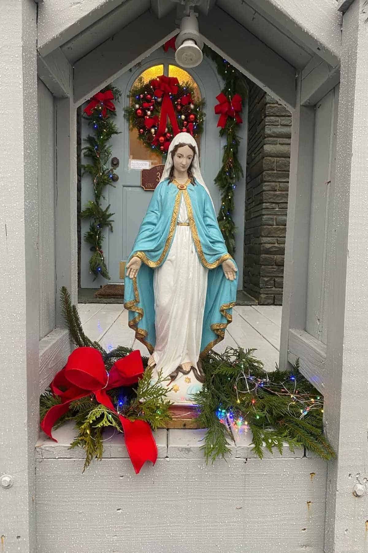 A statue of the Virgin Mary surrounded by greenery, a red ribbon, and Christmas lights, with a decorated door and wreath in the background.