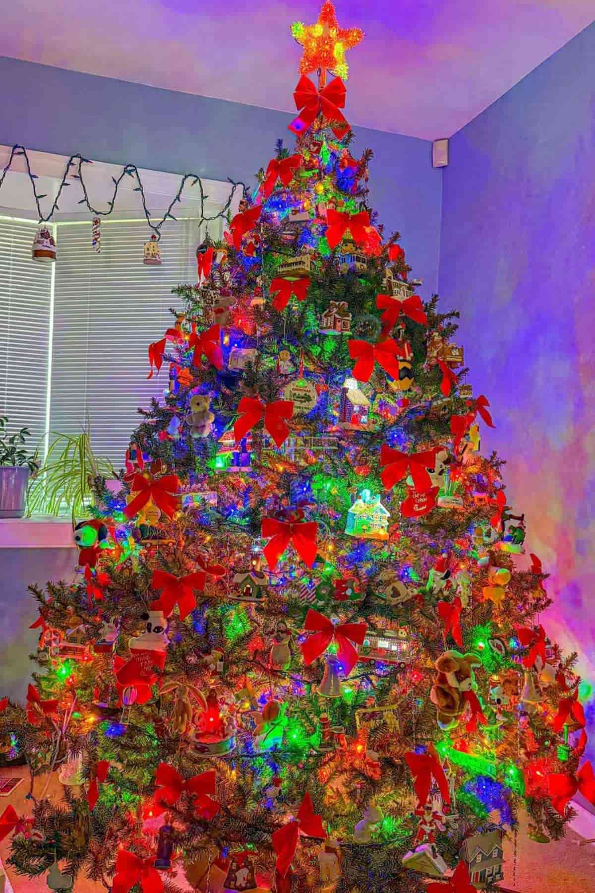 A decorated Christmas tree with red bows, colorful lights, and ornaments stands in a room with blue walls and a window in the background.