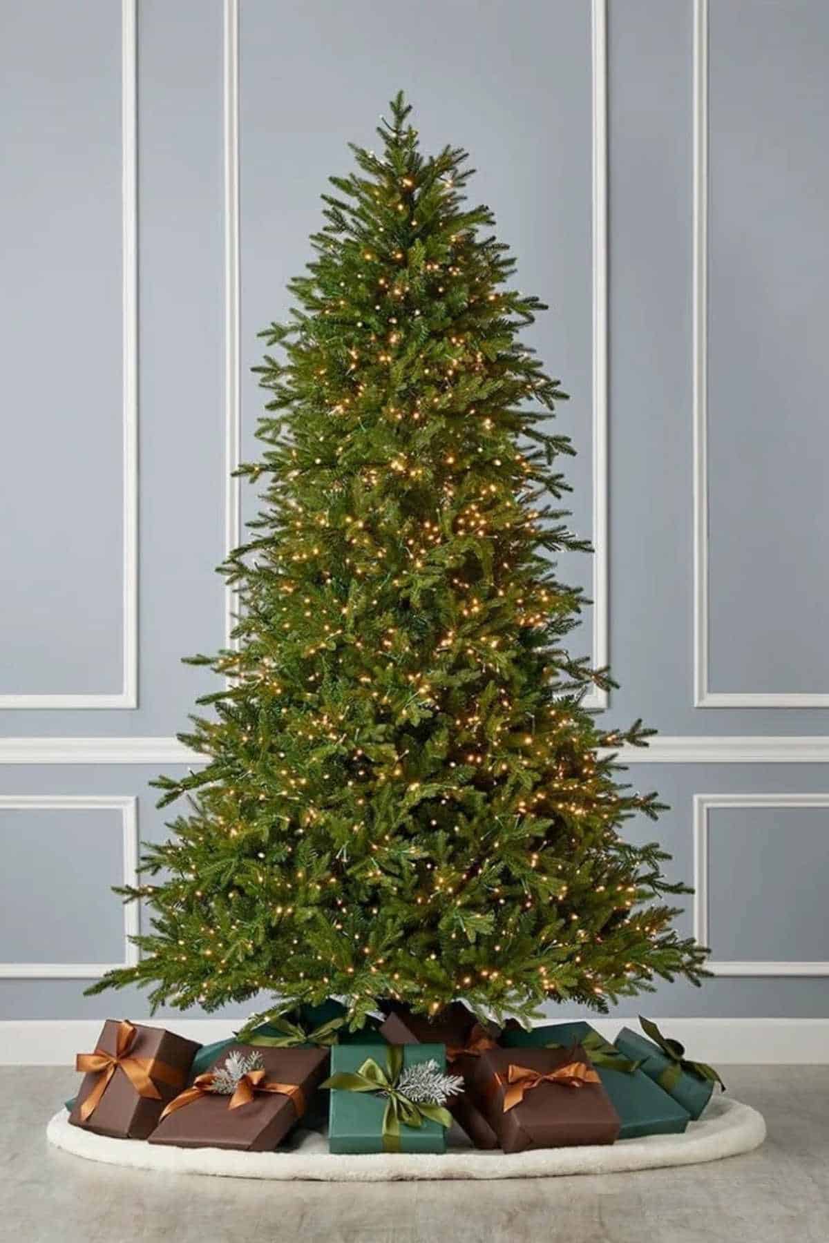 A decorated Christmas tree with white lights stands in front of a gray paneled wall, with wrapped gifts in brown and teal paper placed on a white tree skirt below.