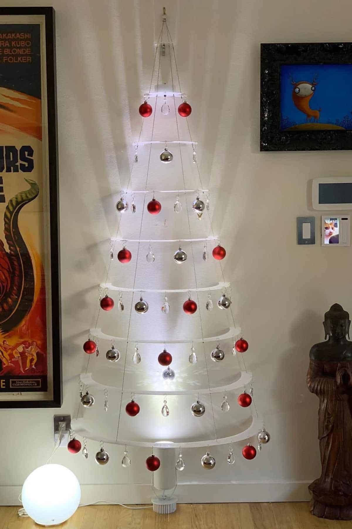 Minimalist Christmas tree made of white wires adorned with red and silver ornaments, illuminated from below, standing against a wall with various decorations.