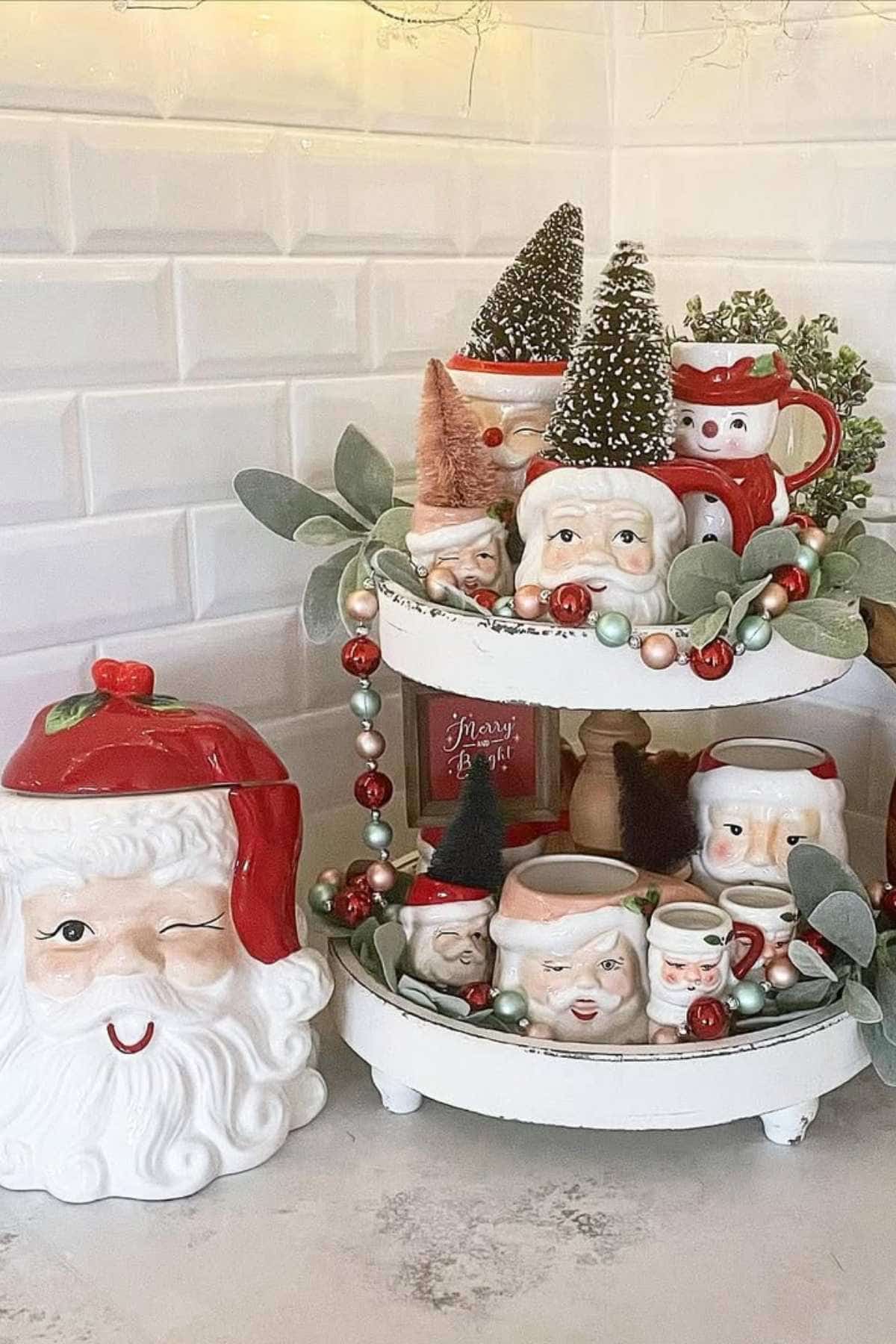 A tiered tray displays vintage Santa mugs, small Christmas trees, faux greenery, and ornaments next to a large winking Santa cookie jar on a kitchen counter.
