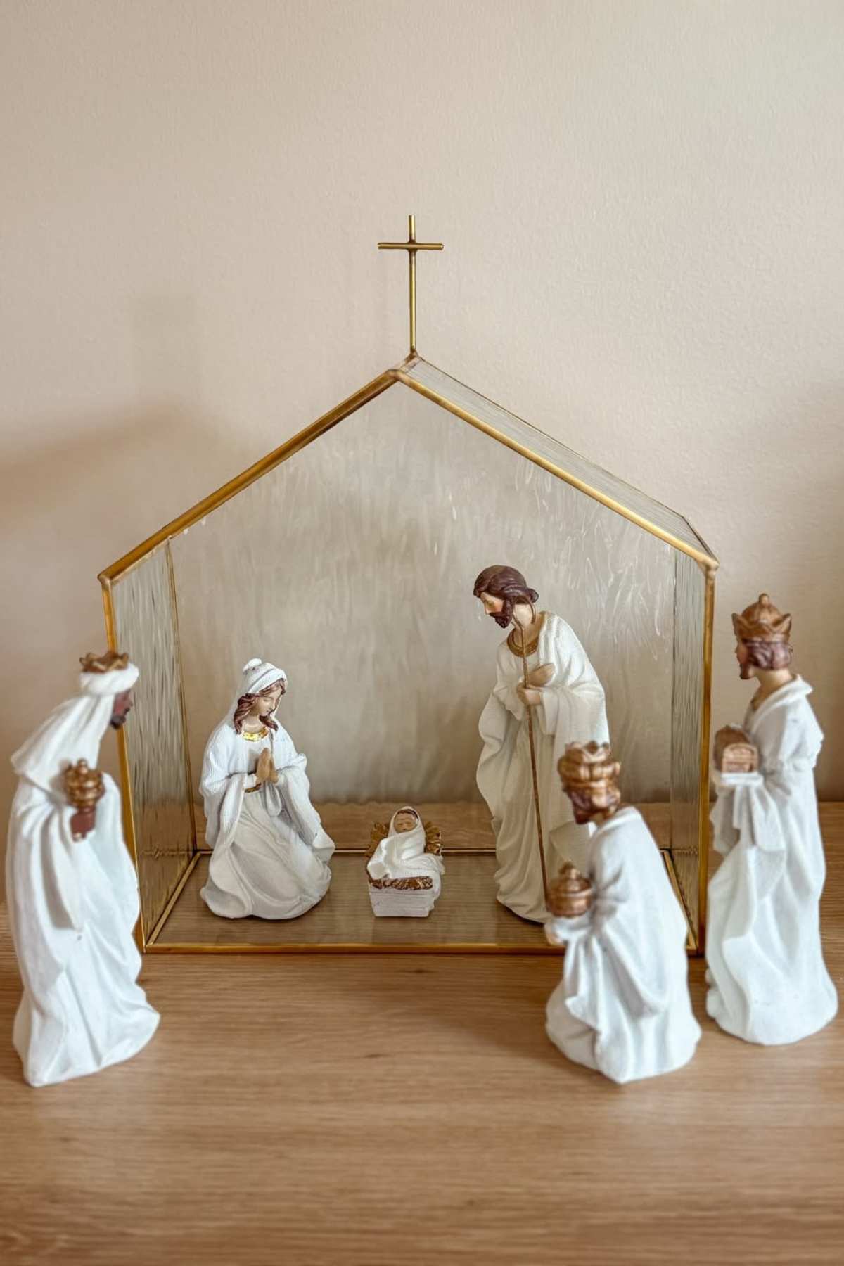 A nativity scene with ceramic figures of Mary, Joseph, baby Jesus in a manger, and three wise men holding gifts, set inside a gold-framed stable on a wooden surface.