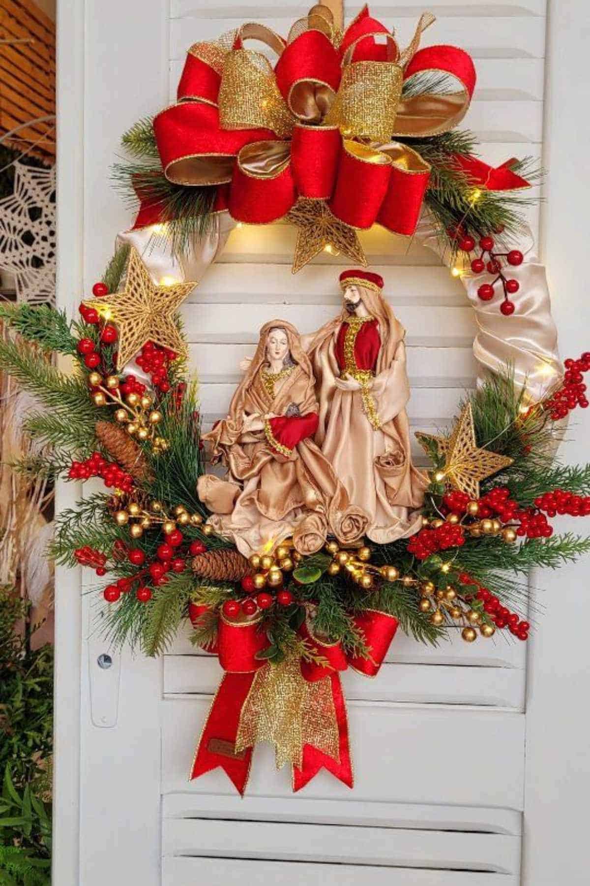 A Christmas wreath decorated with red and gold ribbons, artificial pine branches, berries, and a nativity scene featuring Mary and Joseph in the center.
