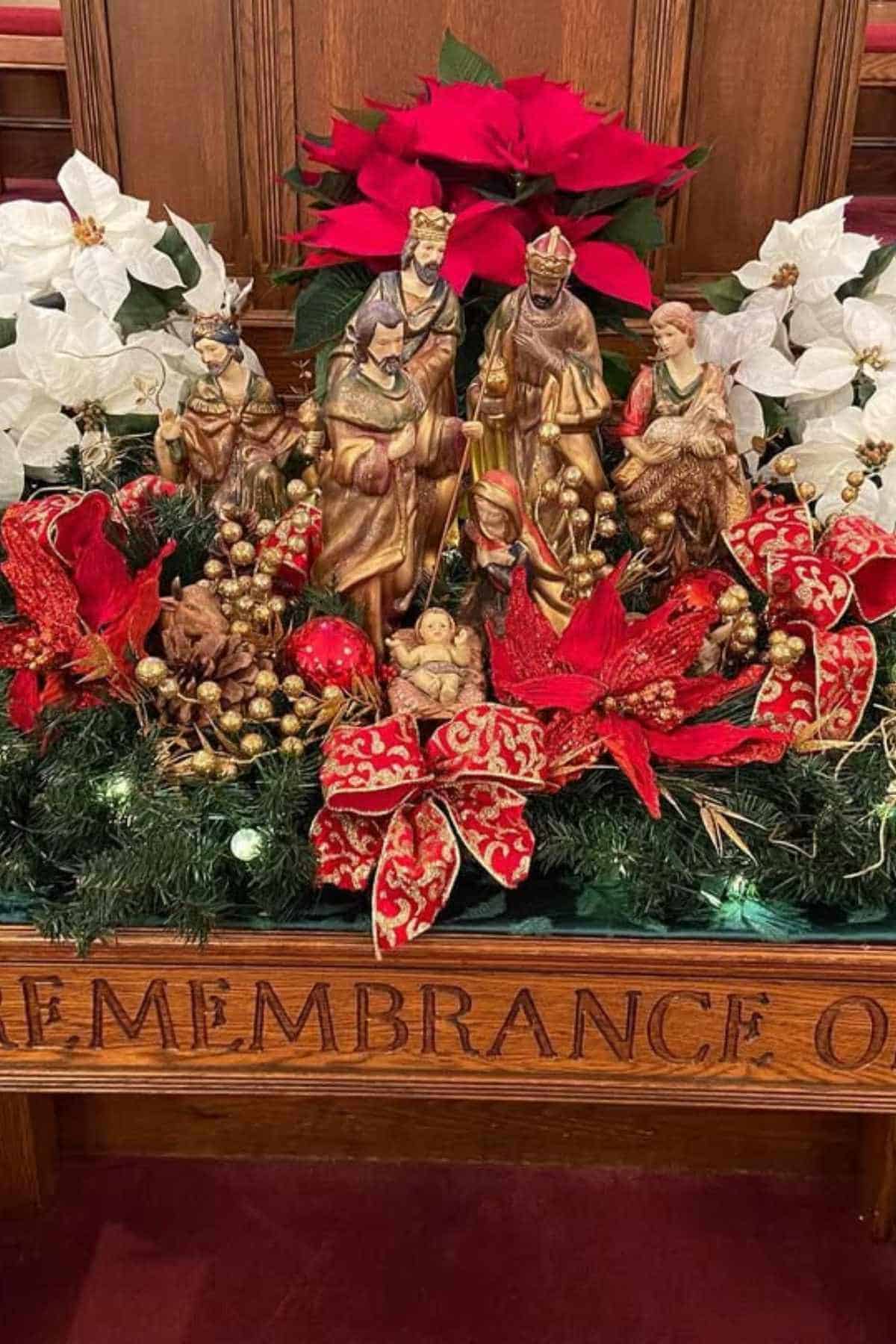 A nativity scene display with figurines, red and white poinsettias, decorative ribbons, and greenery on a wooden table engraved with the word "REMEMBRANCE.