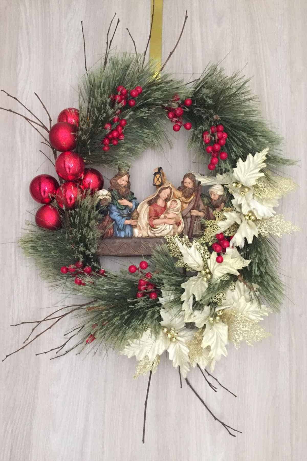 A Christmas wreath decorated with red ornaments, white poinsettias, green pine branches, and a nativity scene in the center featuring baby Jesus, Mary, Joseph, and two others.