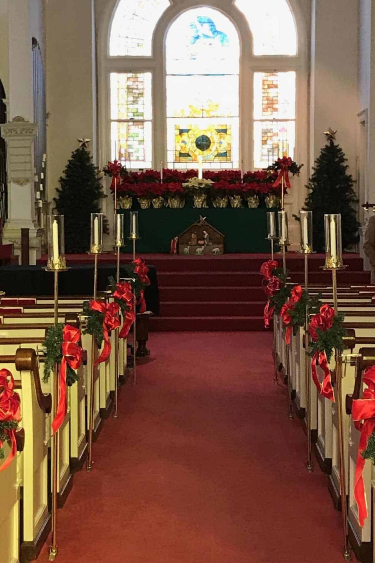 A church sanctuary decorated for Christmas with red bows, candles along the aisle, poinsettias near the altar, two Christmas trees, and a nativity scene in front of stained glass windows.
