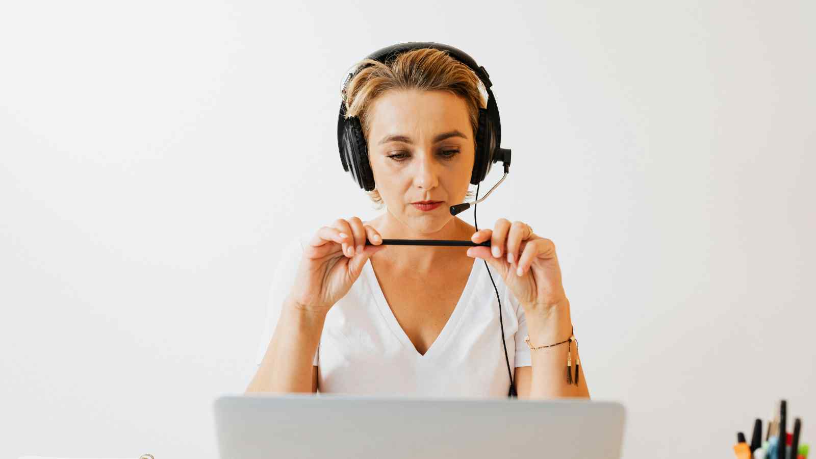 Woman wearing a headset sits at a desk in front of a laptop, holding a pen and appearing focused during a video call or online meeting.