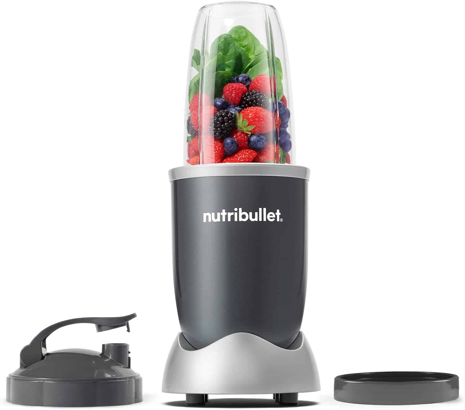 A Nutribullet blender filled with leafy greens and assorted berries, next to a flip-top lid and a flat lid, against a white background.
