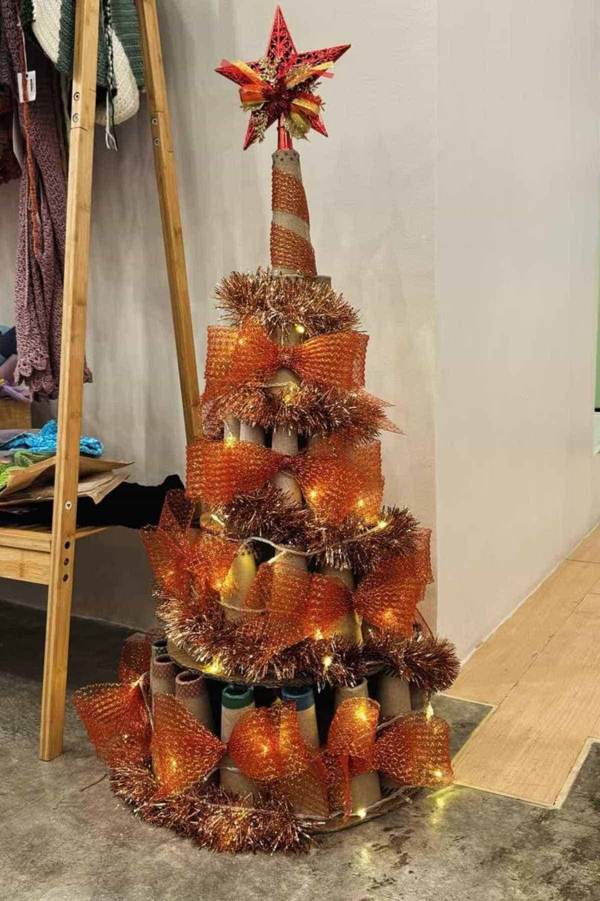 A Christmas tree made from cardboard tubes, orange bubble wrap, tinsel, and string lights, topped with a red star, stands against a wall near a wooden rack with clothes.