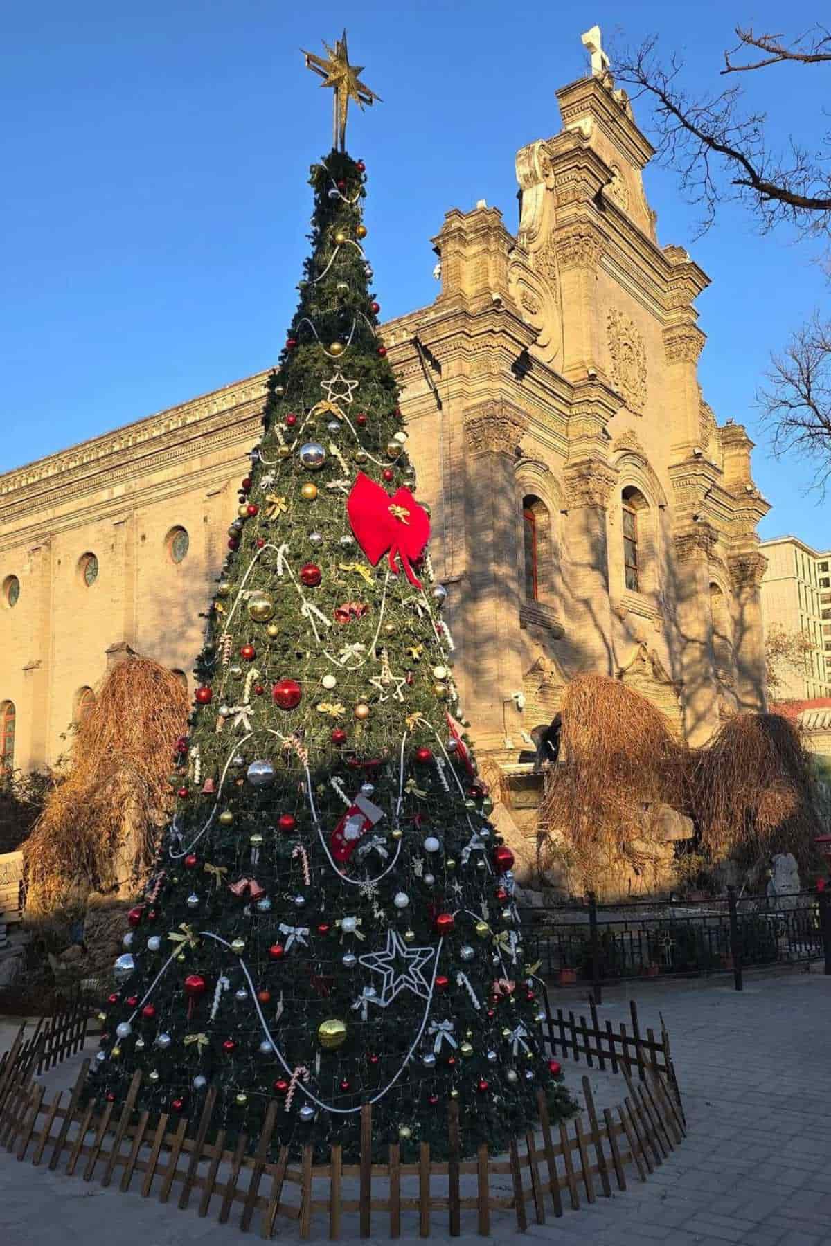 A large decorated Christmas tree with ornaments and a red bow stands in front of a historic stone church on a sunny day.