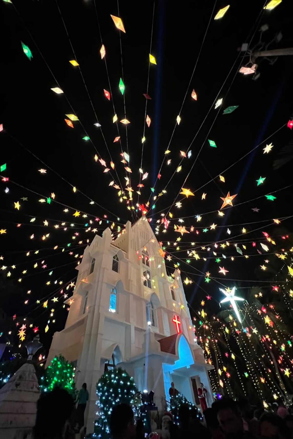 A white church decorated with colorful star-shaped lights overhead at night, with a glowing red cross and illuminated trees and a large star nearby.