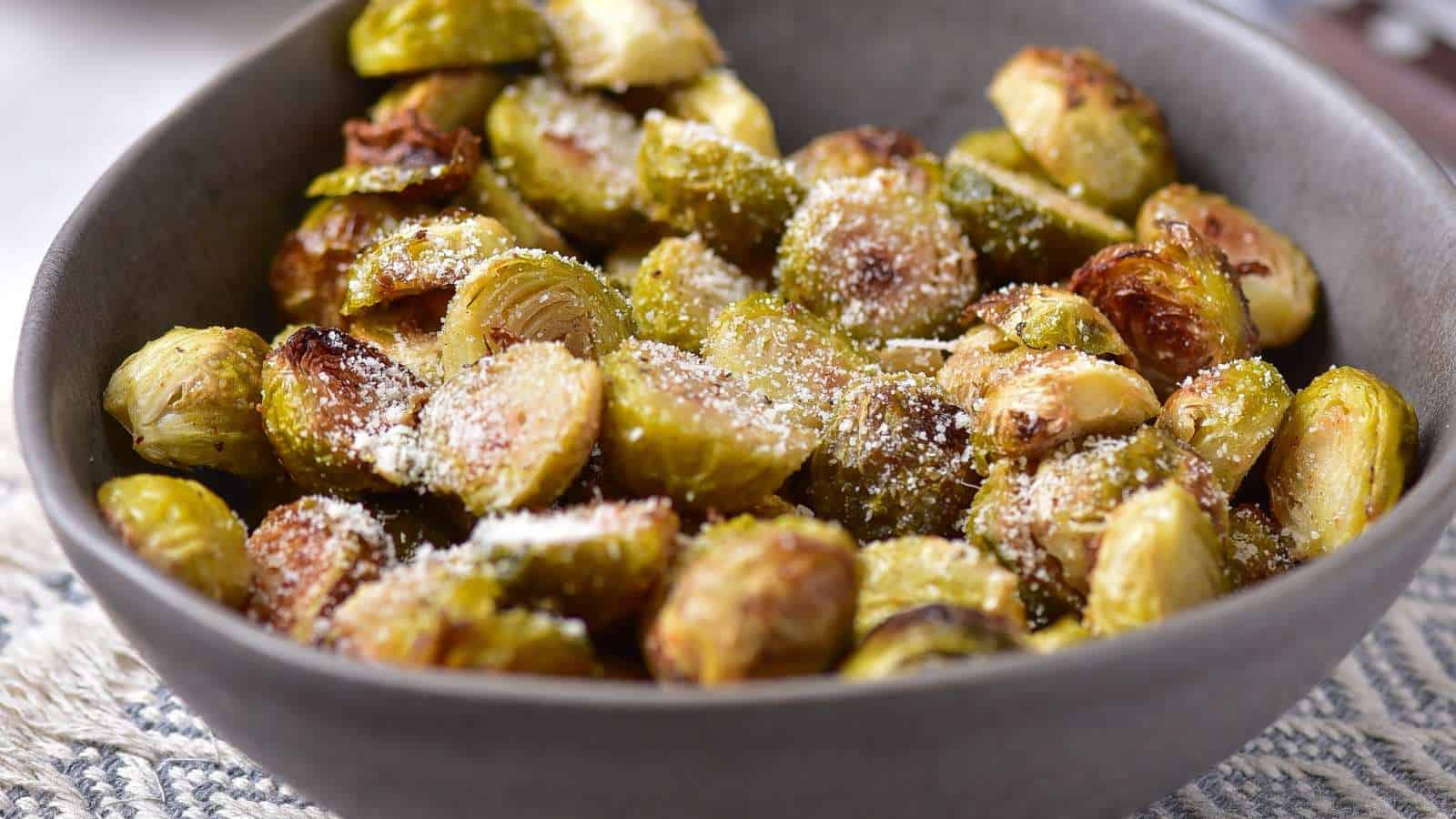 A gray bowl filled with roasted Brussels sprouts sprinkled with grated cheese sits on a patterned tablecloth.