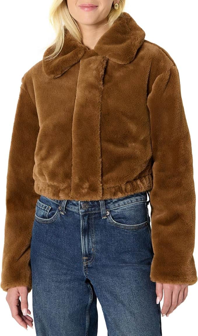 A person wearing a brown faux fur jacket with a wide collar and blue jeans, standing against a plain white background.
