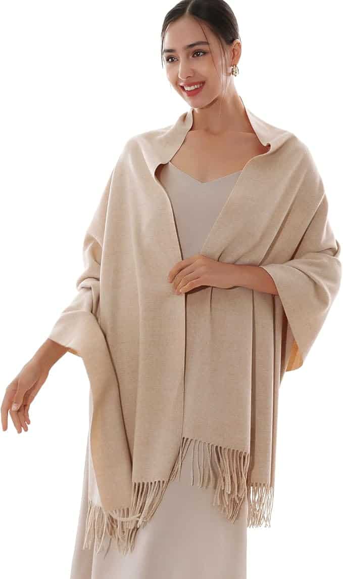 A woman wearing a light beige shawl with fringed edges over a light-colored dress, standing and smiling.