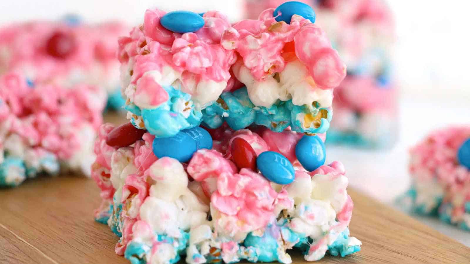Two square popcorn bars with pink, blue, and white colors, topped with red and blue candy-coated chocolates, are stacked on a wooden surface.