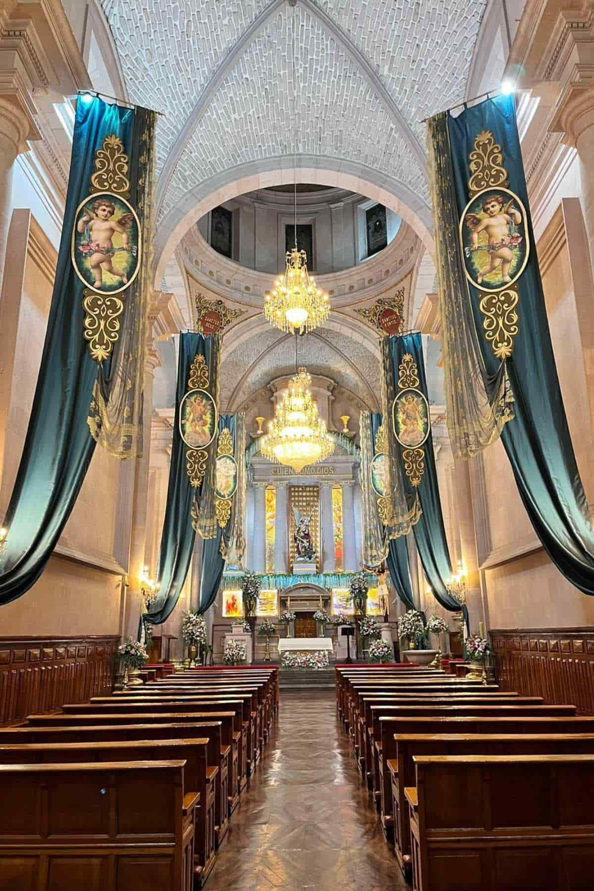 Interior of a church with chandeliers, tall ceilings, wooden pews, ornate banners, and an altar decorated with religious artwork and floral arrangements.