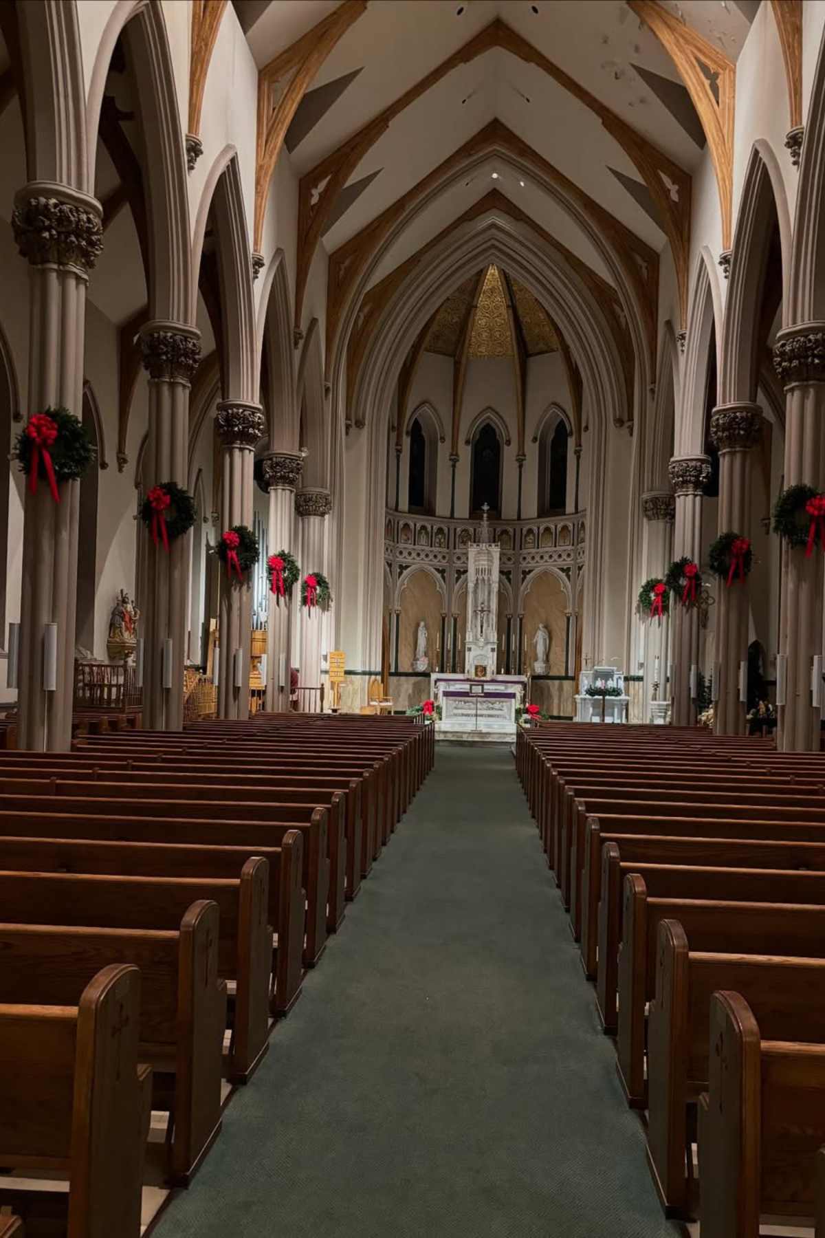 Interior view of a church with wooden pews, vaulted ceiling, columns, altar at the front, and holiday wreaths hanging on pillars.