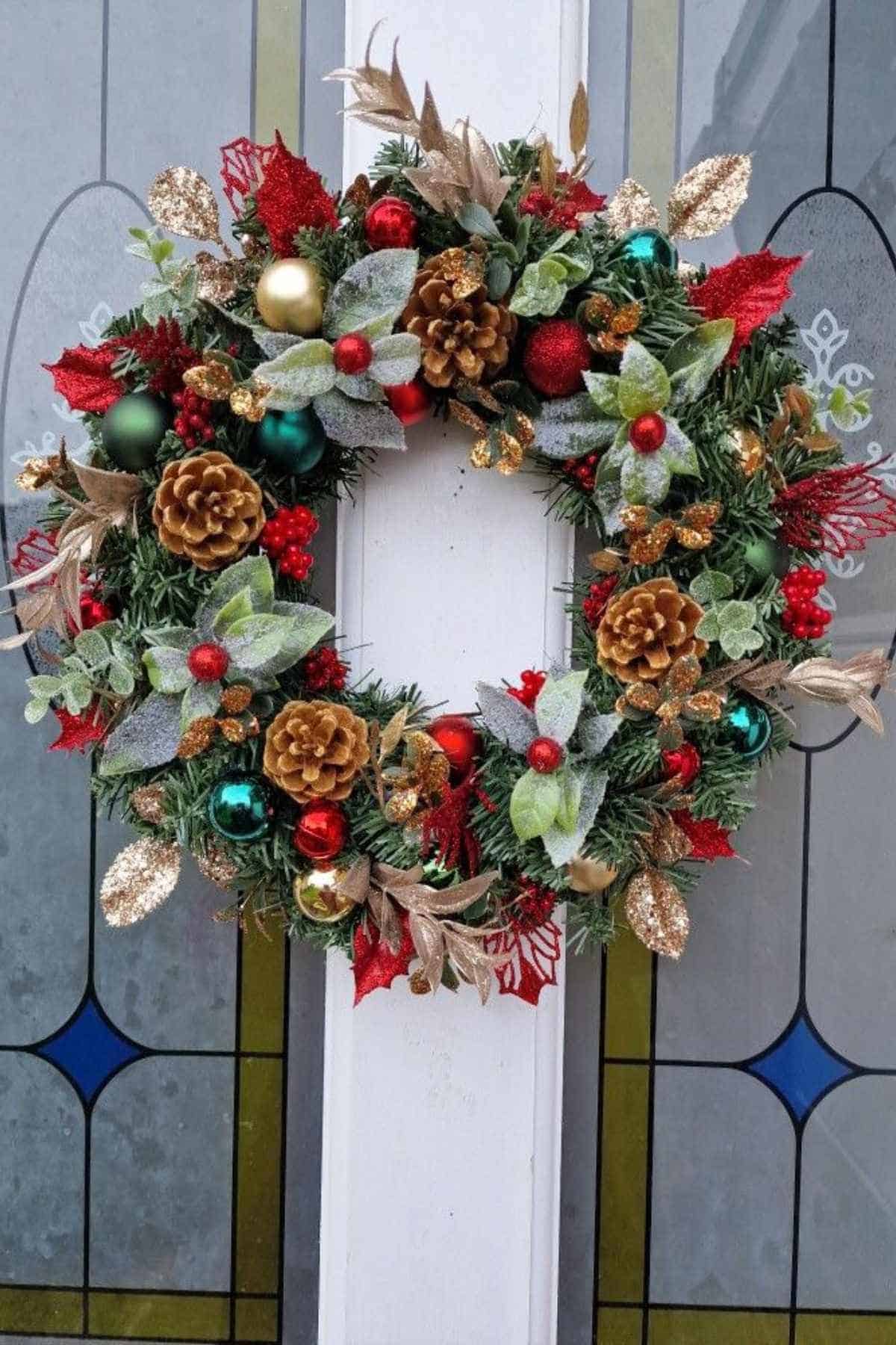 A festive Christmas wreath with pine cones, red berries, gold leaves, and colorful ornaments hangs on a glass-paneled door.