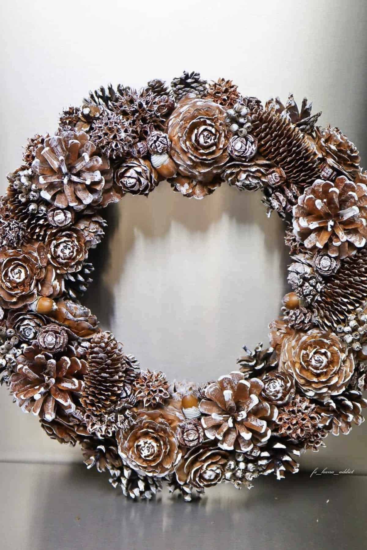 A decorative wreath made of pinecones and small berries, lightly dusted with white to resemble snow, displayed against a plain background.