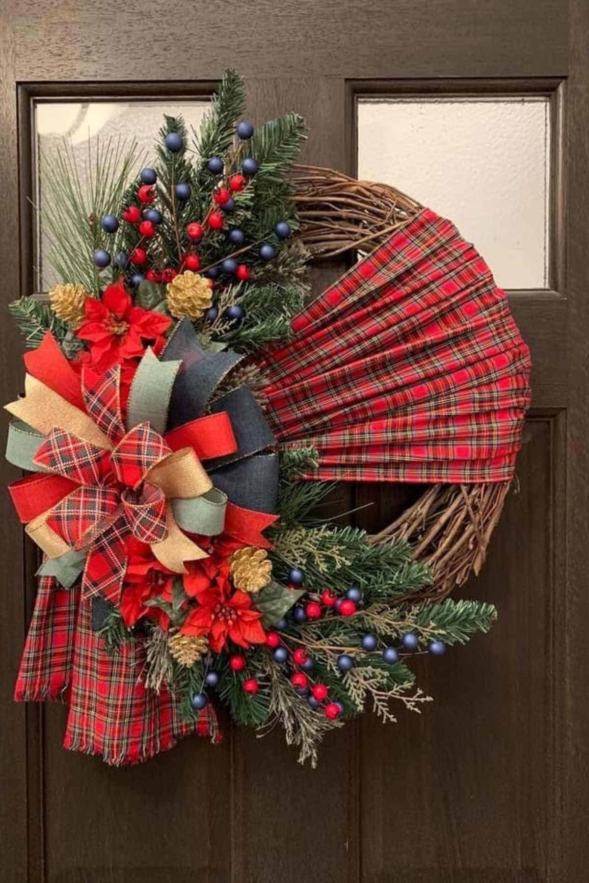 A holiday wreath on a dark door featuring red plaid fabric, artificial pine branches, red berries, blue berries, gold pinecones, and a plaid bow.