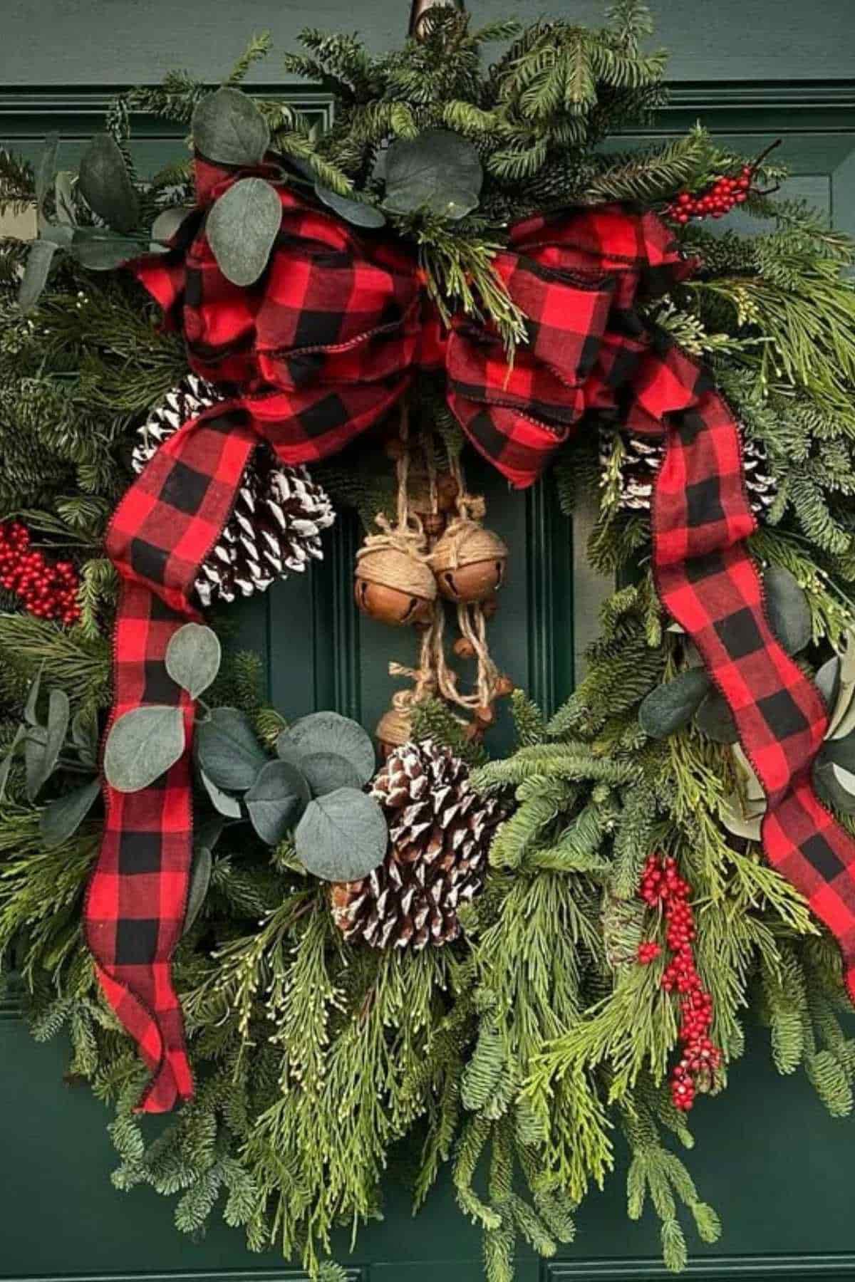A holiday wreath with pine branches, red berries, pinecones, eucalyptus leaves, a red-and-black plaid bow, and hanging jingle bells, displayed on a green door.