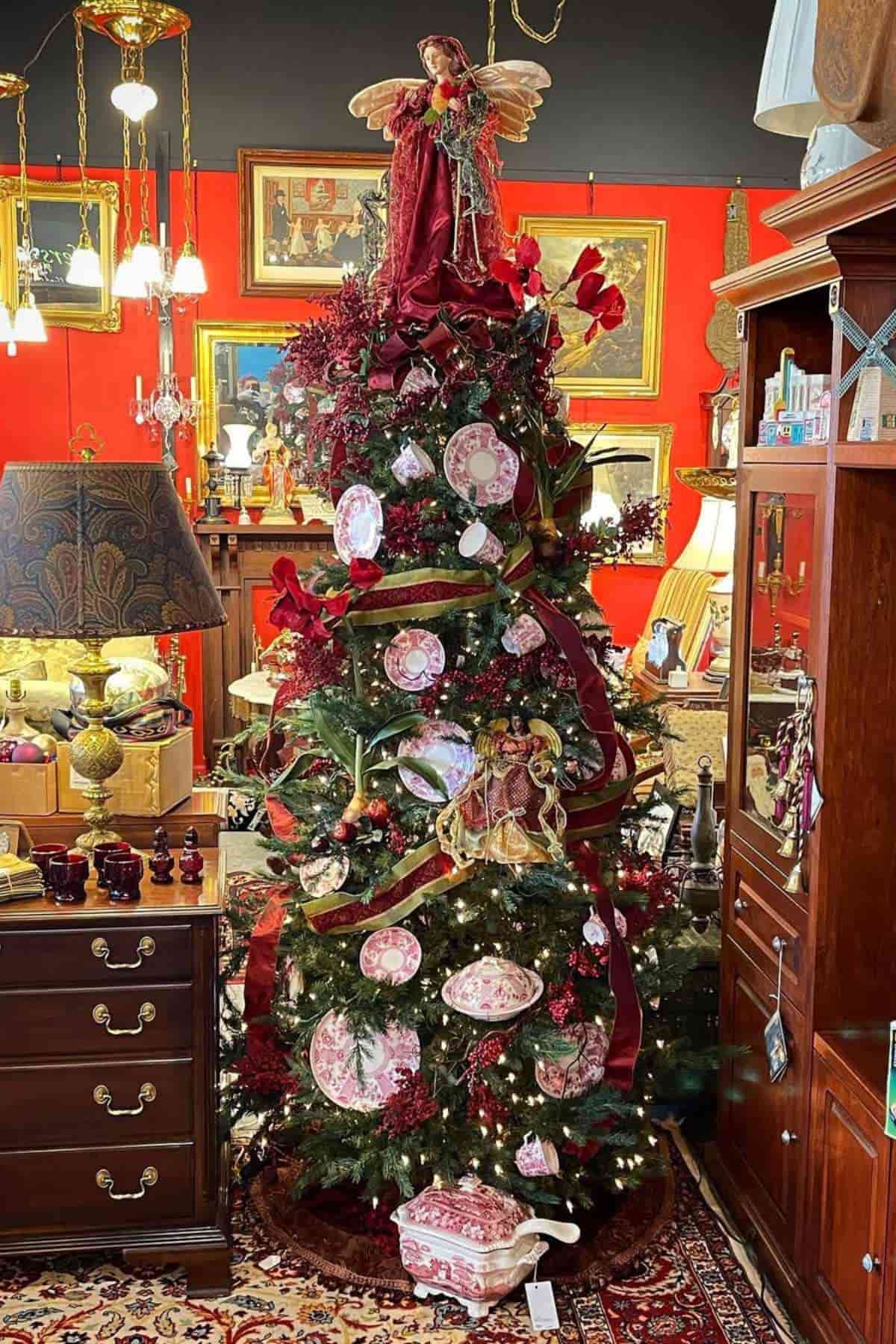 A decorated Christmas tree in a vintage room, topped with an angel and adorned with red ornaments and decorative plates, surrounded by antique furniture and framed artwork.