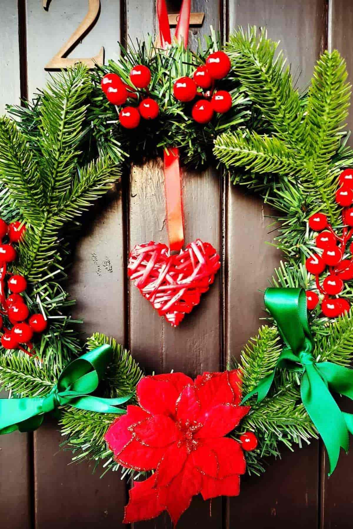 A festive wreath with green pine needles, red berries, a red heart ornament, a red poinsettia flower, and green ribbons hangs on a dark wooden door.
