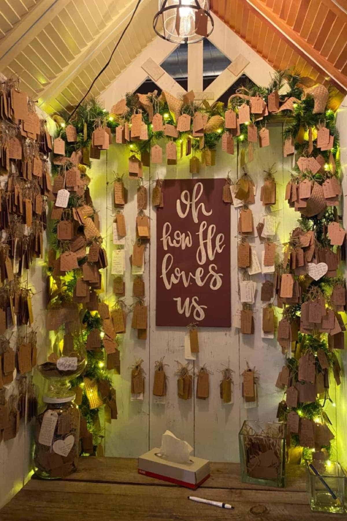 Small wooden room decorated with string lights, greenery, and many handwritten tags. A large sign in the center reads, “Oh how He loves us.” A tissue box sits on the table below.