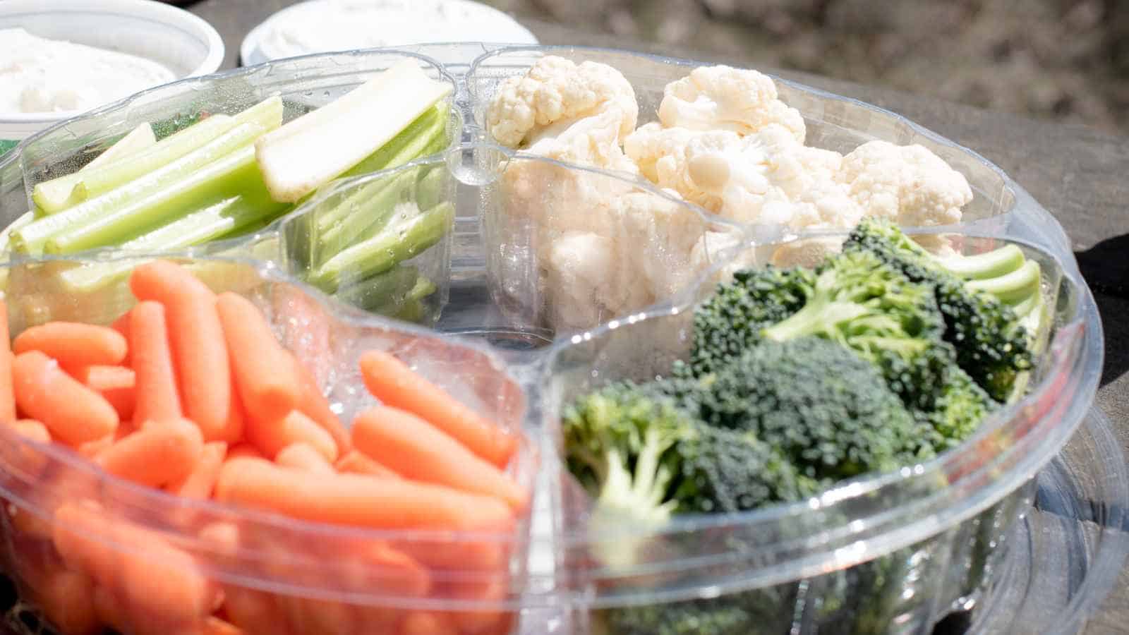 A plastic tray with sections containing raw celery sticks, cauliflower florets, baby carrots, and broccoli florets.