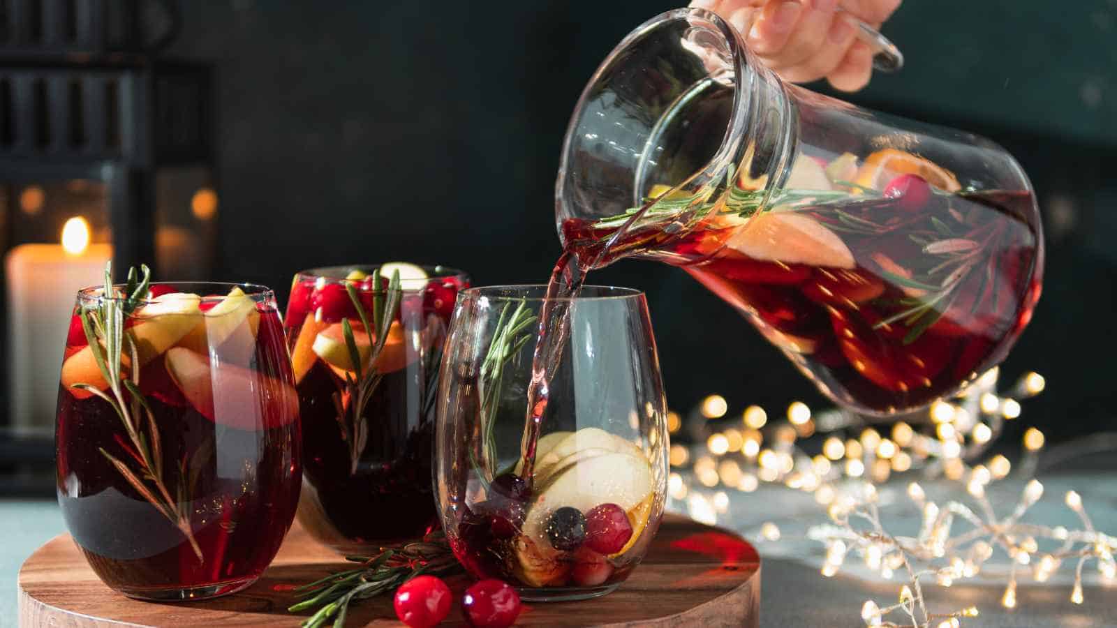 A hand pours red sangria with fruit and rosemary from a glass pitcher into a glass beside two filled glasses, with a candle and string lights in the background.
