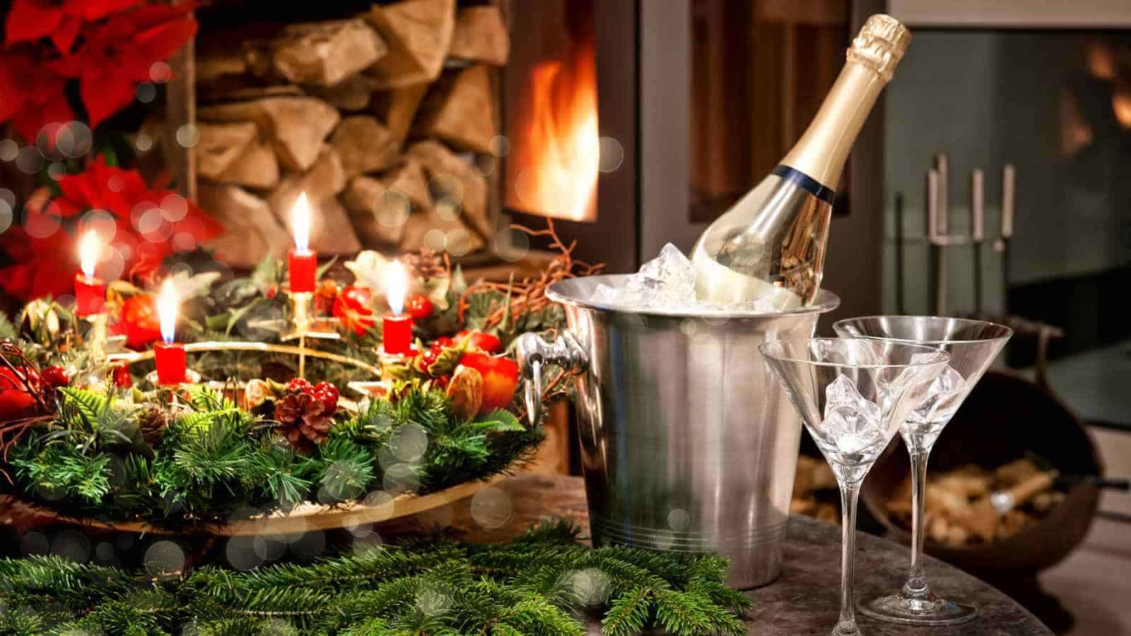 A bottle of champagne in an ice bucket, two glasses with ice, and a festive wreath with lit red candles are on a table near a fireplace.