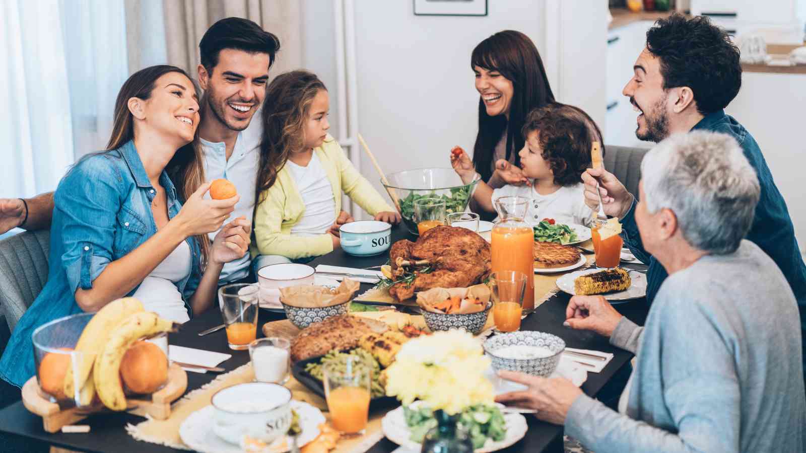 A group of people of various ages sit around a table, sharing a meal with roasted turkey, fruit, and drinks, smiling and enjoying each other's company.