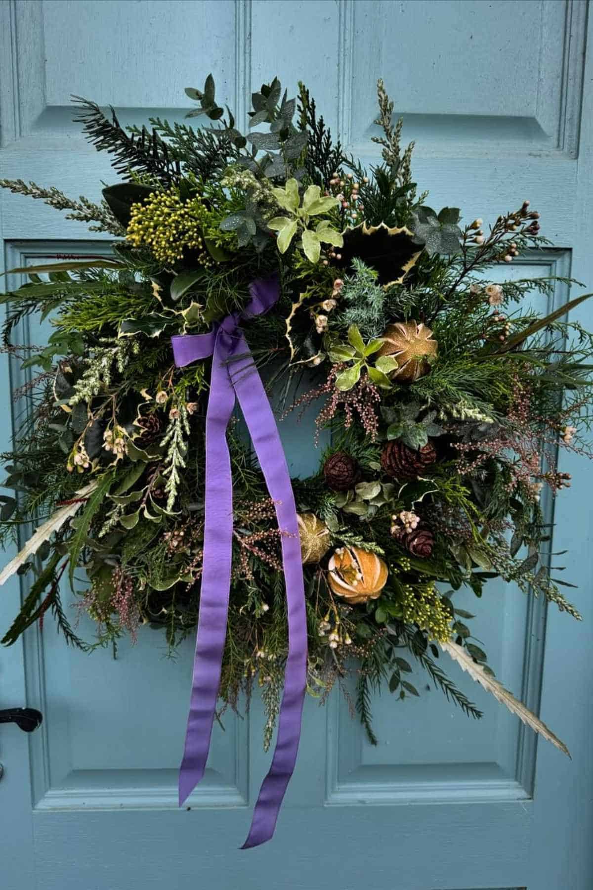 A decorative wreath made of green foliage, dried flowers, and pinecones hangs on a blue door, adorned with a long purple ribbon.