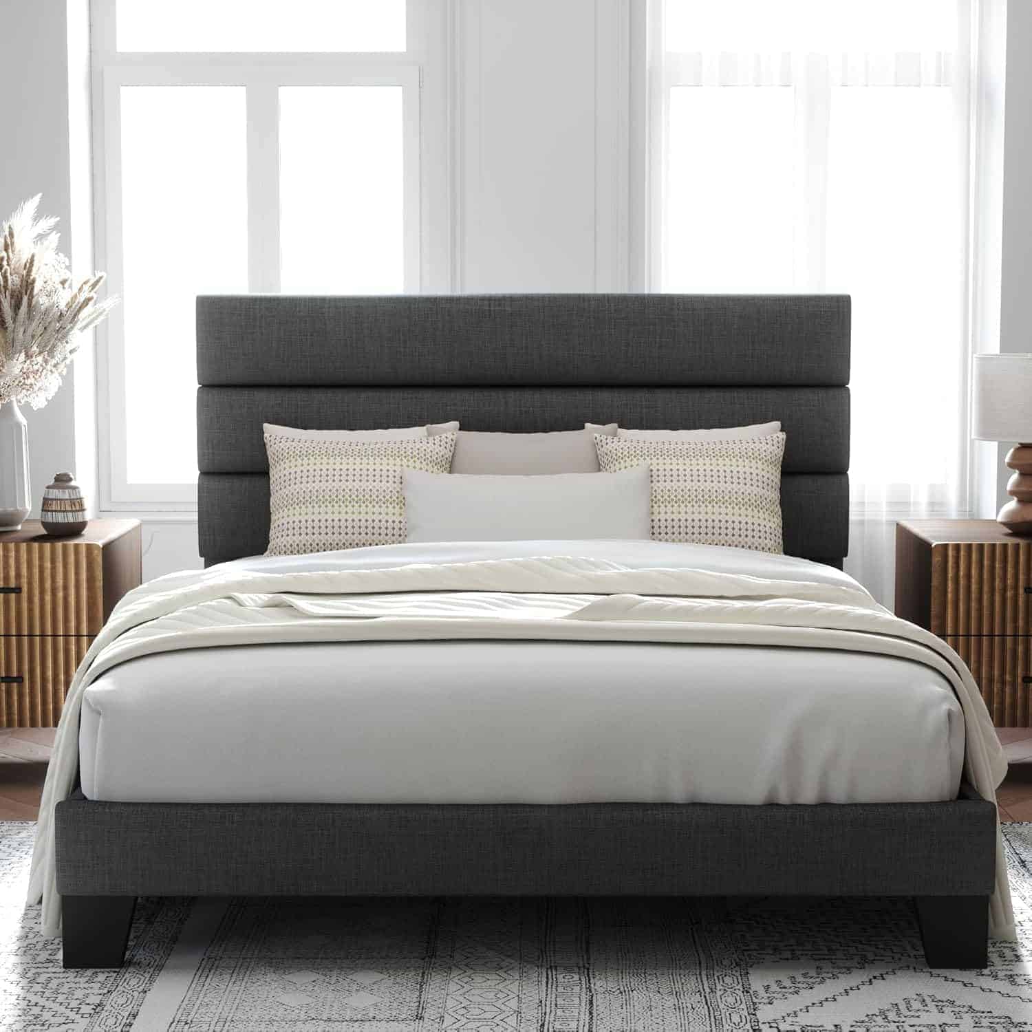 A modern bedroom with a dark gray upholstered bed, neutral bedding, striped pillows, and matching wood nightstands with lamps, set against large windows.
