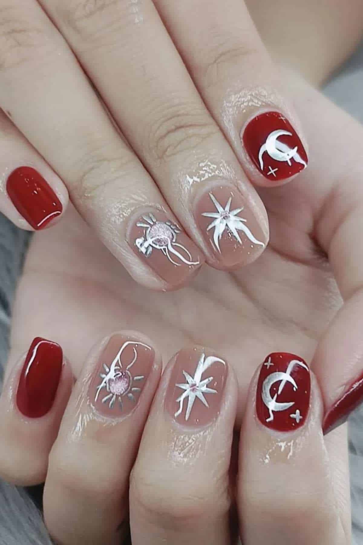 A hand with red and nude-colored nails featuring white celestial designs, including stars, the moon, and planets.
