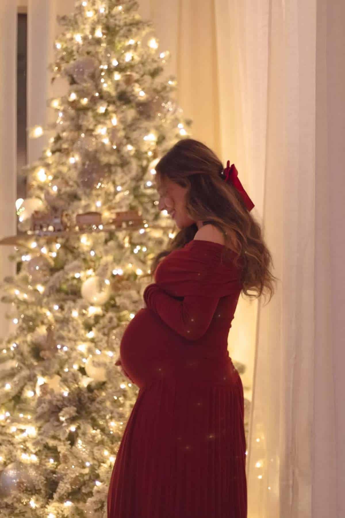 A pregnant woman in a red dress stands in front of a decorated, lit Christmas tree indoors.