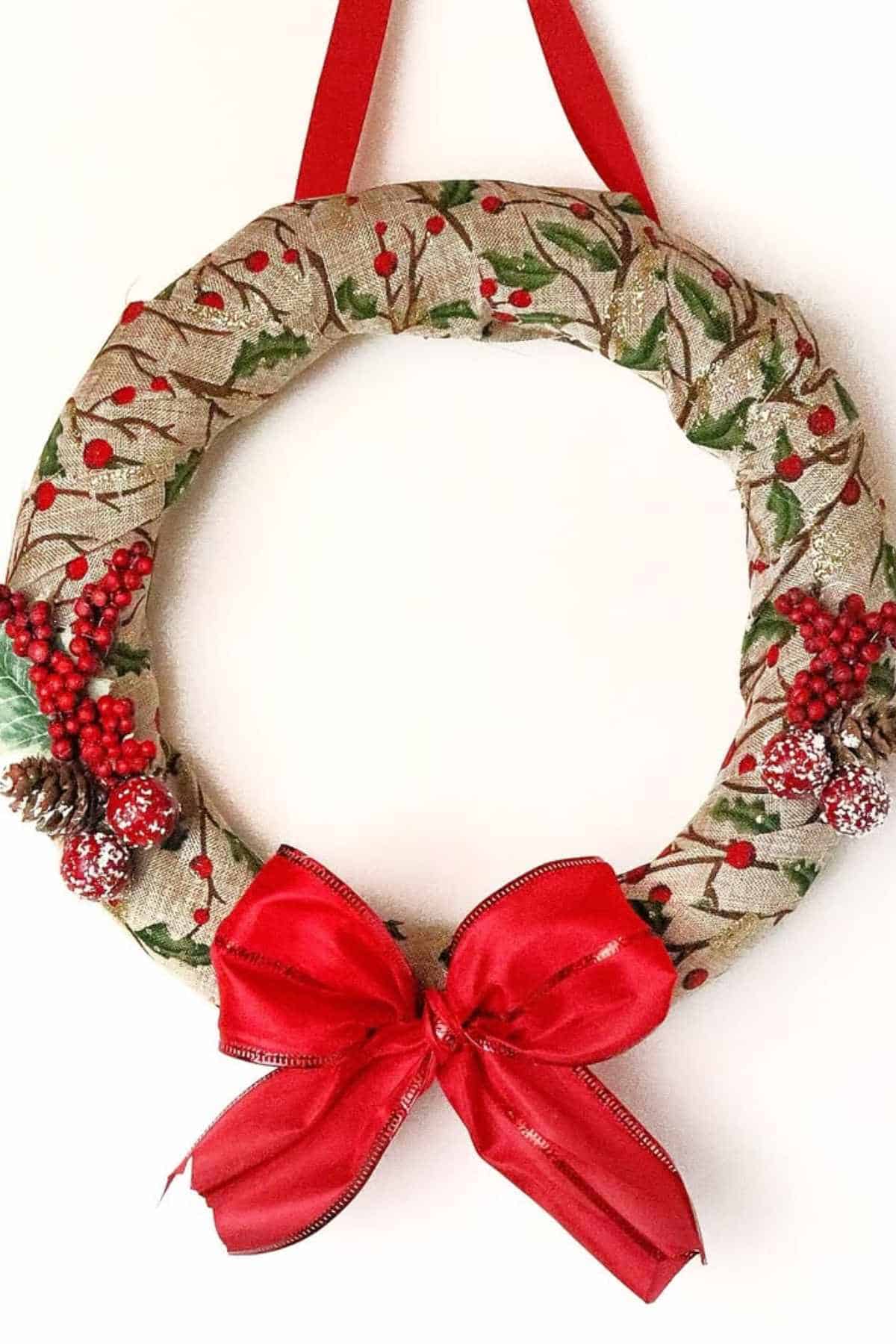 A decorative wreath wrapped in fabric with holly and berry patterns, adorned with red berries, pine cones, and a large red bow, hangs from a red ribbon.