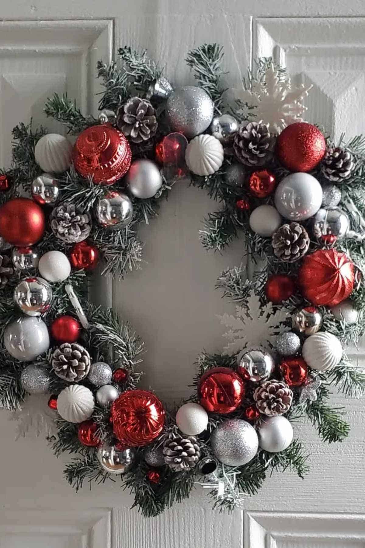 A holiday wreath decorated with red, silver, and white ornaments, pinecones, and artificial greenery hangs on a white door.