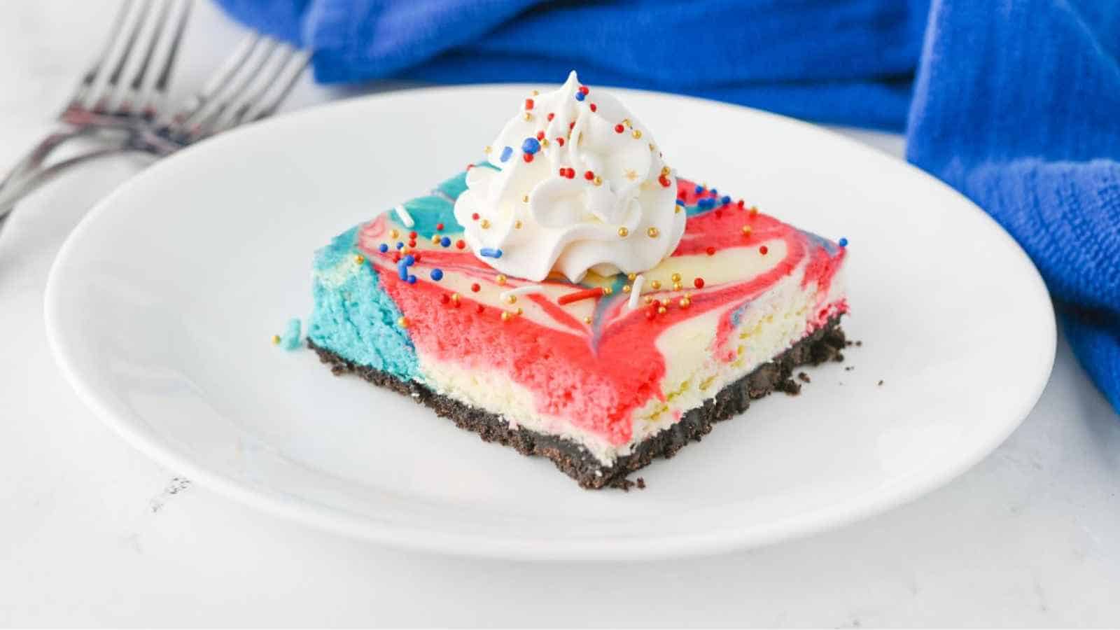 A square slice of colorful red, blue, and white cheesecake with whipped cream and sprinkles on top, served on a white plate.