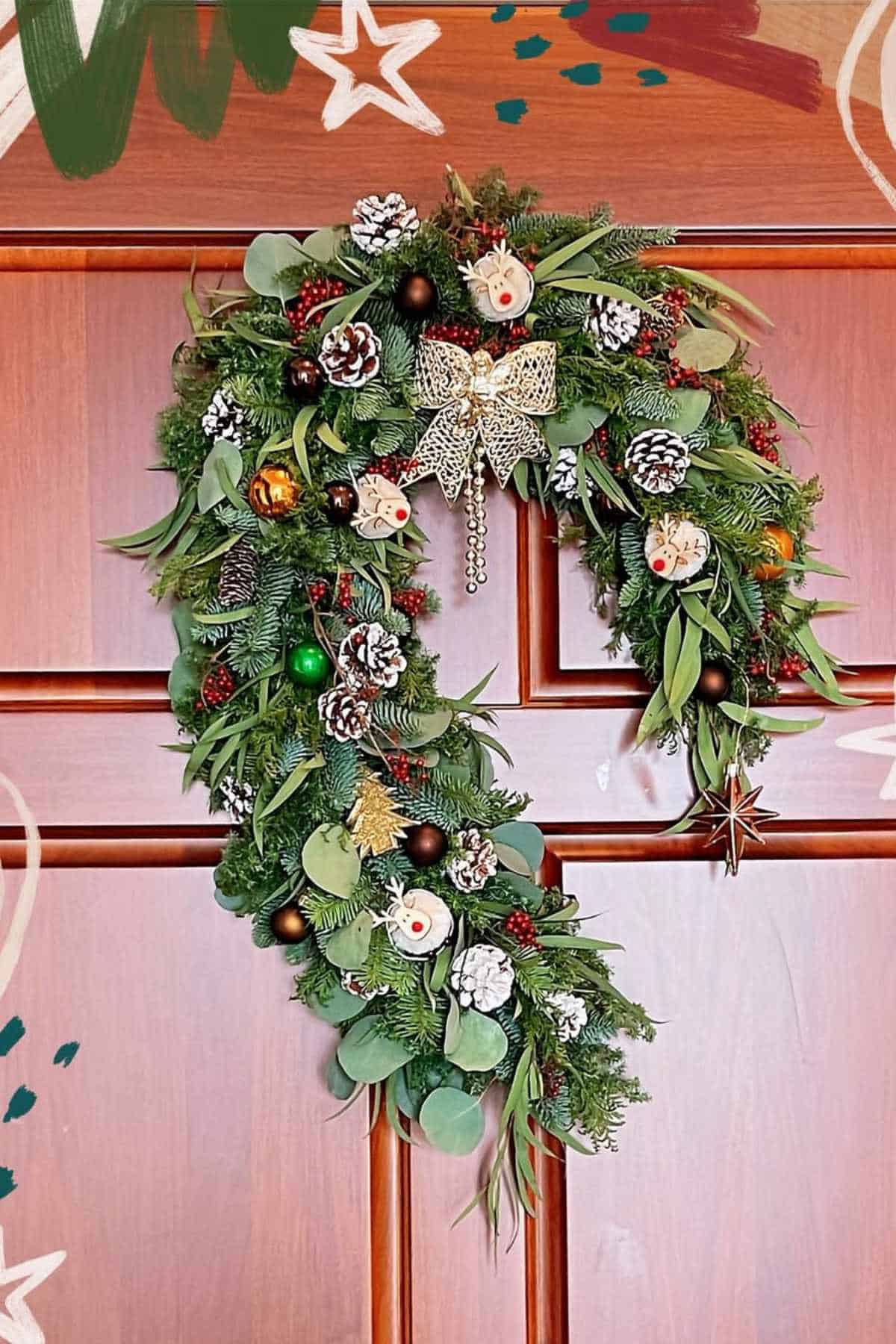 A Christmas wreath shaped like a candy cane, decorated with pinecones, ornaments, greenery, a bow, and faux berries, hangs on a wooden door.
