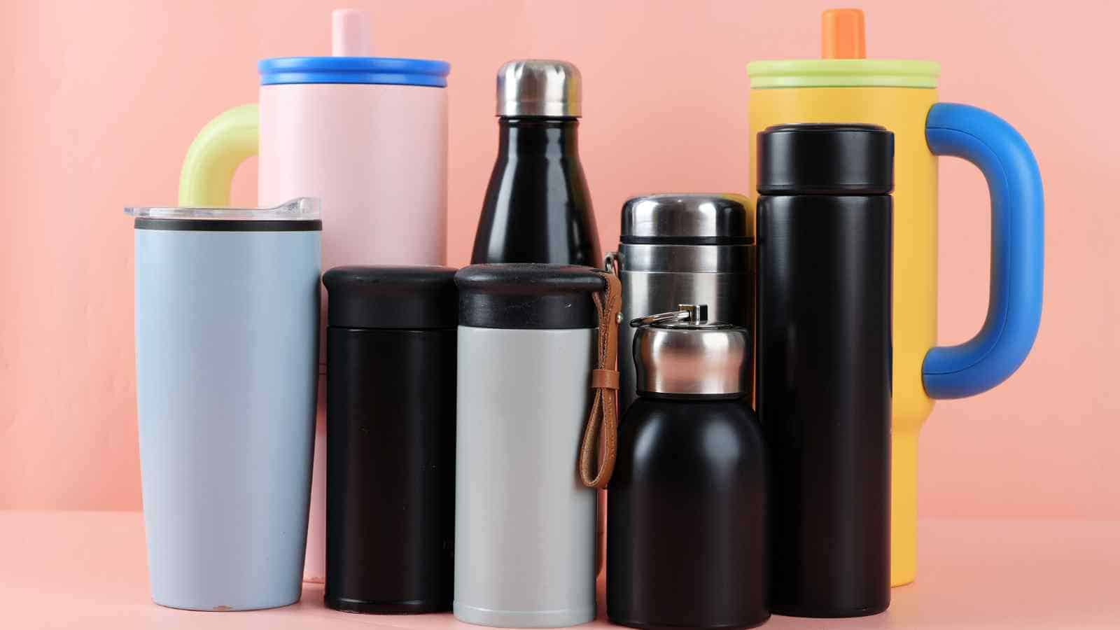 A group of reusable travel mugs and water bottles in various colors, shapes, and sizes arranged against a pink background.