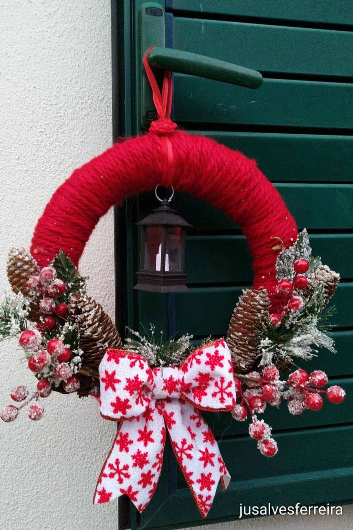 A red yarn-wrapped wreath with pine cones, red berries, a lantern, and a white bow with red snowflakes hangs on a door handle.