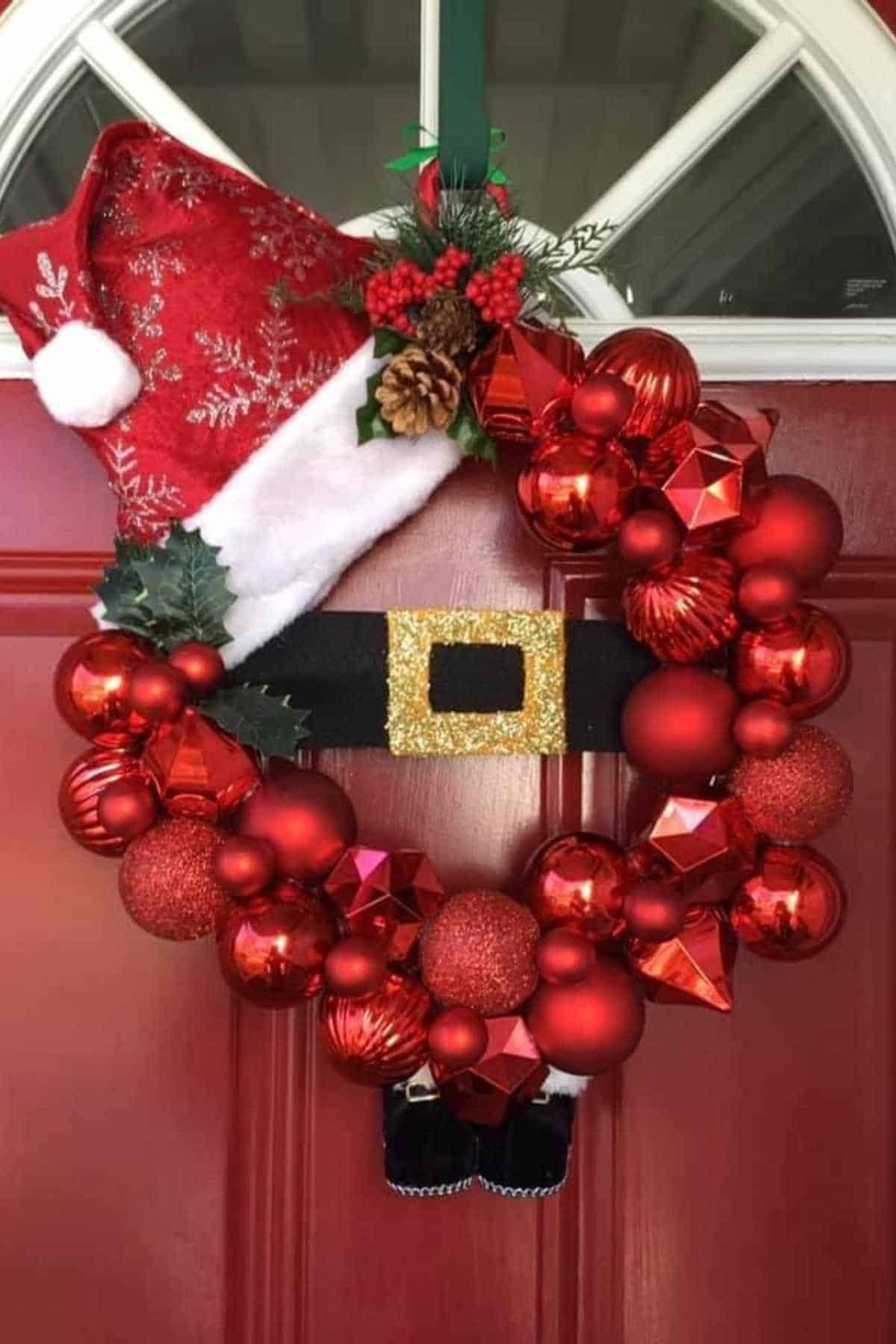 A Christmas wreath made of red ornaments with a Santa hat, black belt, gold buckle, and small boots hangs on a red door.