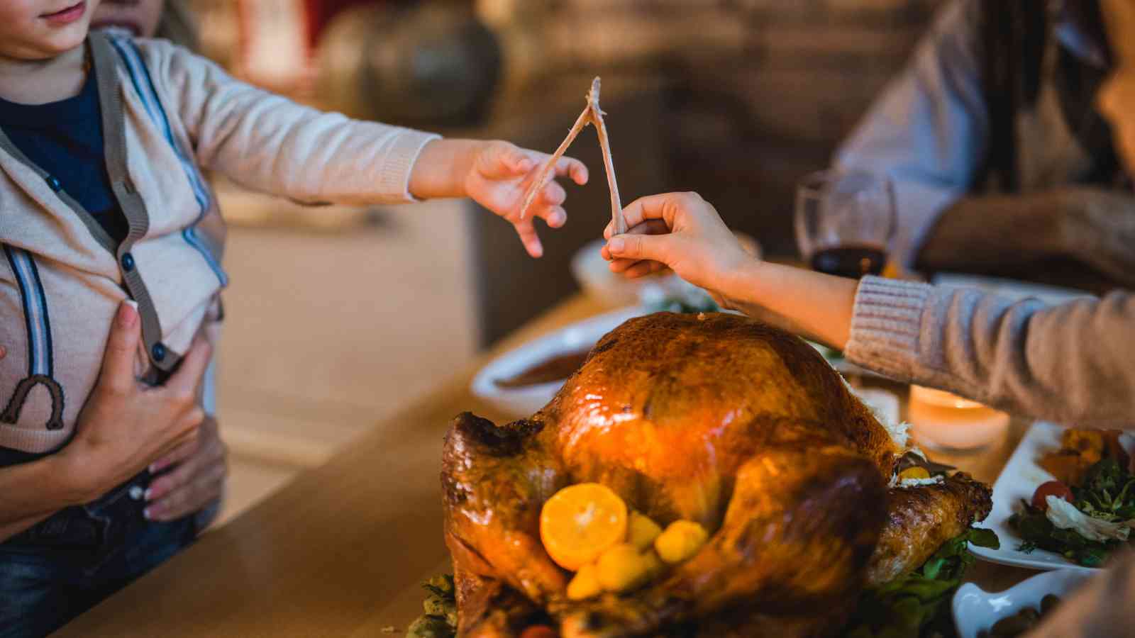 Two people hold opposite ends of a wishbone above a roasted turkey on a dining table set with food.