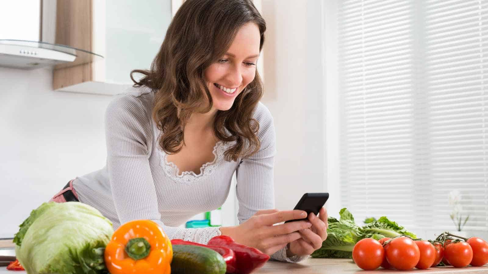 Woman in a kitchen smiling at her phone, surrounded by fresh vegetables including lettuce, bell pepper, tomato, and cucumber on the counter.