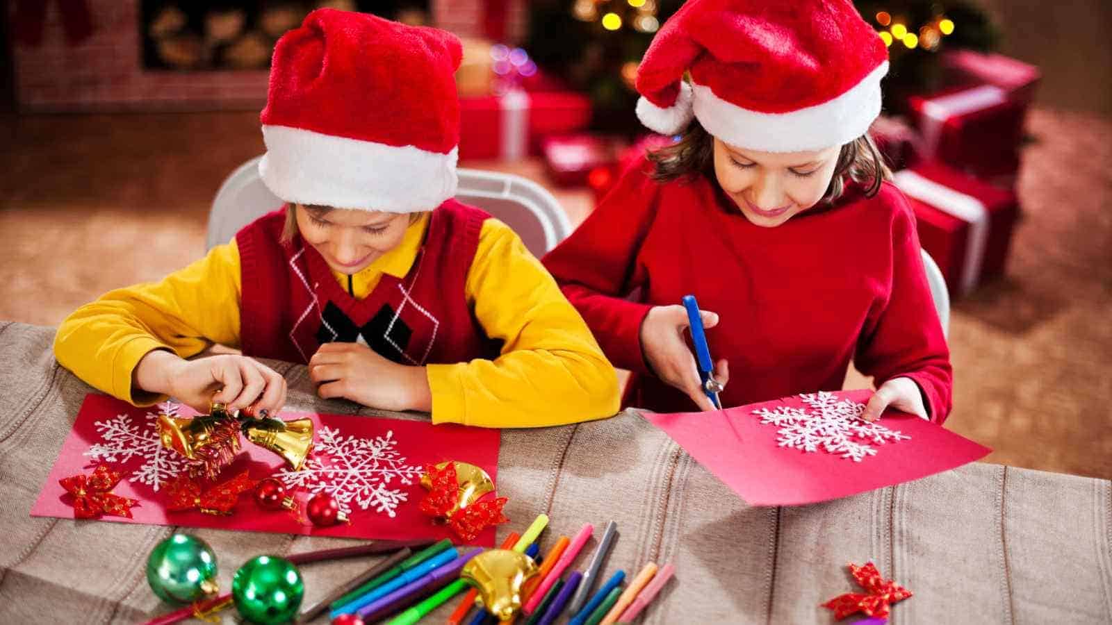Two children wearing Santa hats make Christmas crafts at a table with colored markers, scissors, and decorations.
