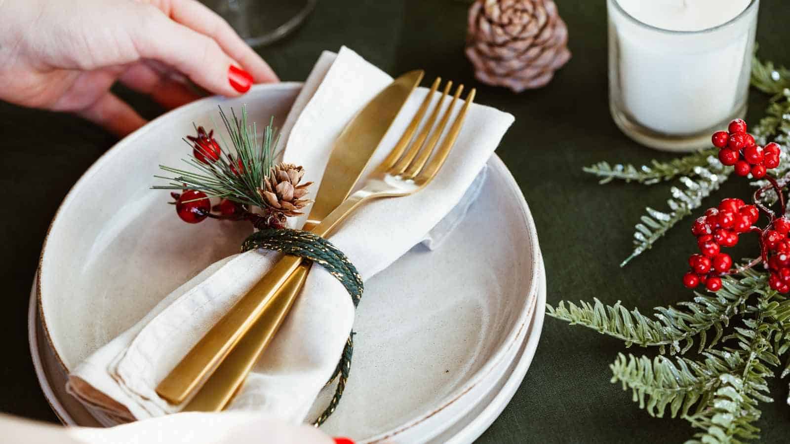 A hand arranges a holiday table setting with gold cutlery wrapped in a napkin, decorated with pine and berries, on stacked plates with festive greenery and a candle nearby.